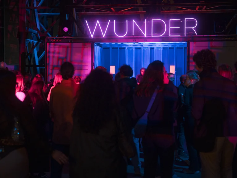 Eine Menschenmenge tanzt vor einem blau und pink beleuchteten Container, in dem eine DJ auflegt. Über dem Container leuchtet der Schriftzug „WUNDER“ in violettem Neonlicht.