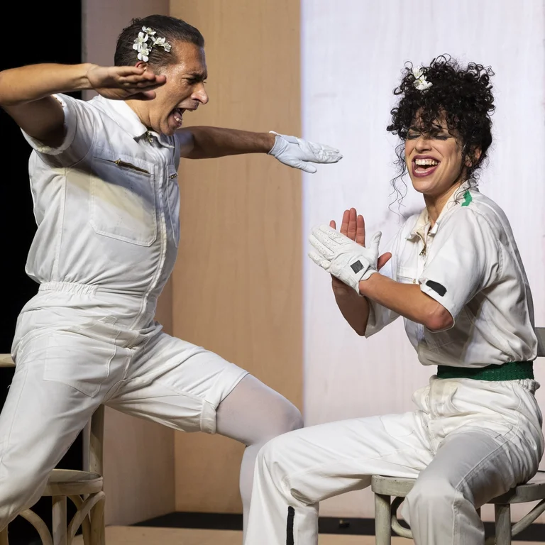 Marlene Monteiro Freitas und Israel Galván stand close to each other in white jumpsuits, both raising their arms to shoulder height.