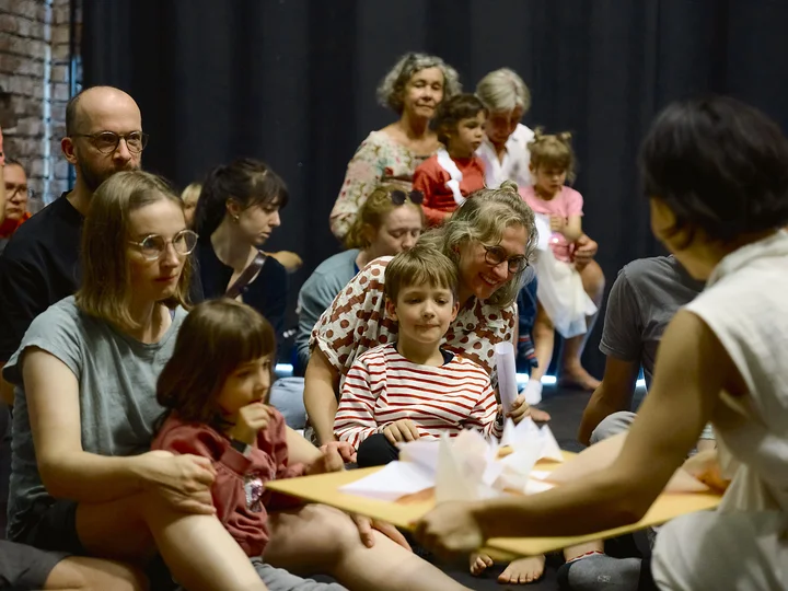 Adults and children sit closely together in a room during the show of "Club Origami" at Ruhrtriennale 2025.