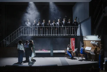 Stage view with a group of uniformed men standing on an elevated platform. Below them are four performers, while three musicians play cello, piano and violin on the right.