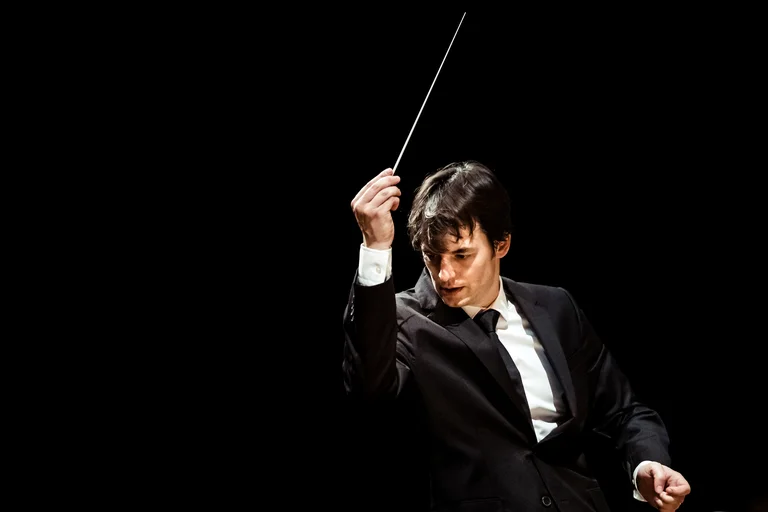 Julien Masmondet stands in a black suit with a white shirt and a black tie against a dark background. With intense focus, he raises the baton into the air.