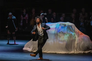 A barefoot performer in a dark suit dances on stage in front of a large draped object illuminated by colorful projections, with other people visible in the dark background.