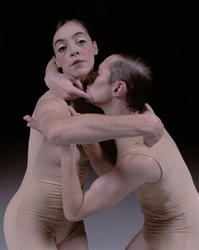 Two dancers in skin-toned costumes stand closely entwined, with one gently holding the other’s head. The scene radiates a blend of intimacy, vulnerability, and quiet intensity.