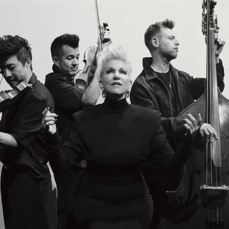 Schwarz-weiß-Aufnahme von Joyce DiDonato, die von den drei Musiker:innen von Time for Three umgeben ist, die auf ihren Streichinstrumenten spielen.