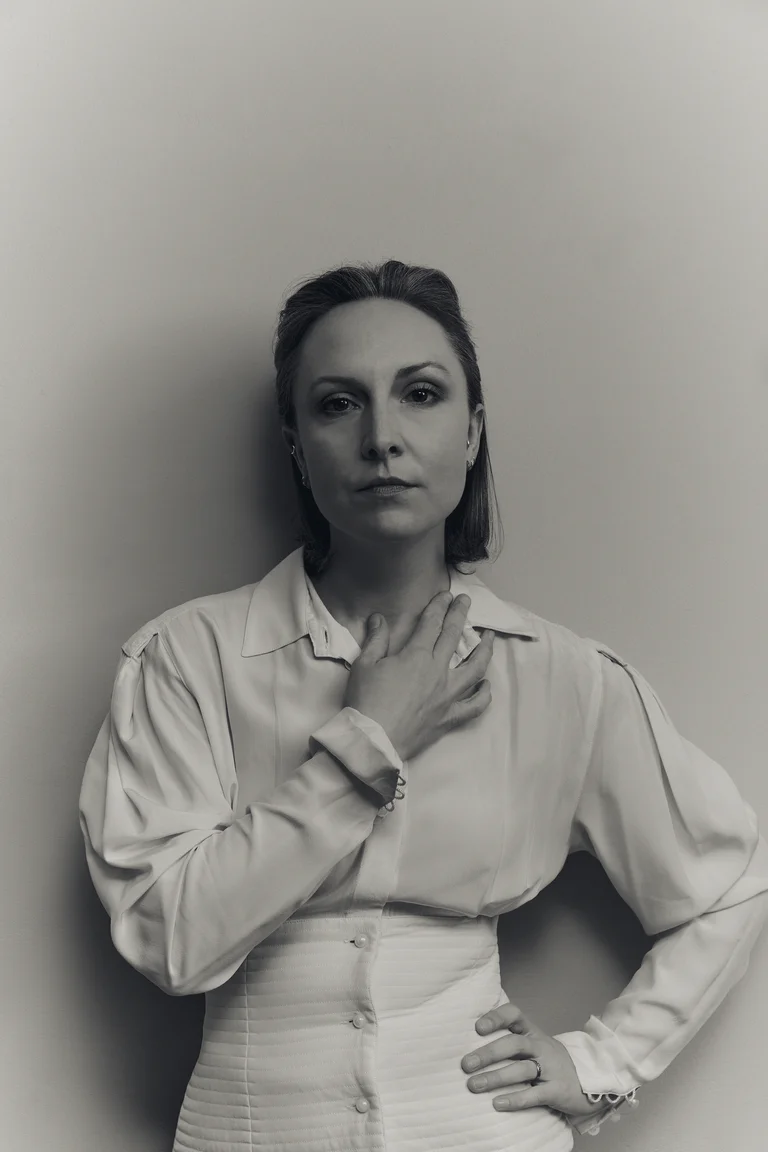Eliza McCarthy stands in front of a plain wall. Her facial expression is serious and calm, her gaze directed straight at the camera. One hand rests on her neck, the other on her hip.