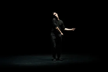A dancer dressed in black stands in the spotlight on a dark stage, performing a restrained movement.