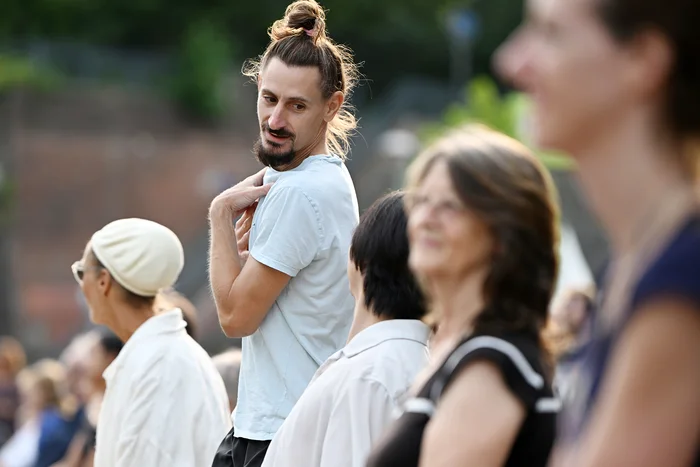Ioannis Mandafounis tanzt in einem weißen Shirt. Etwas verschwommen im Vordergrund sieht man das Publikum wie es den Tänzer:innen gespannt zu guckt