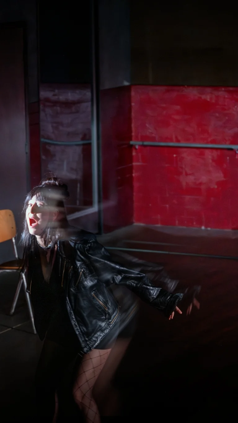 On a stage with red and grey wall surfaces, a person stands in the foreground with arms outstretched. Several chairs are arranged in the space, with focused lighting illuminating the scene.
