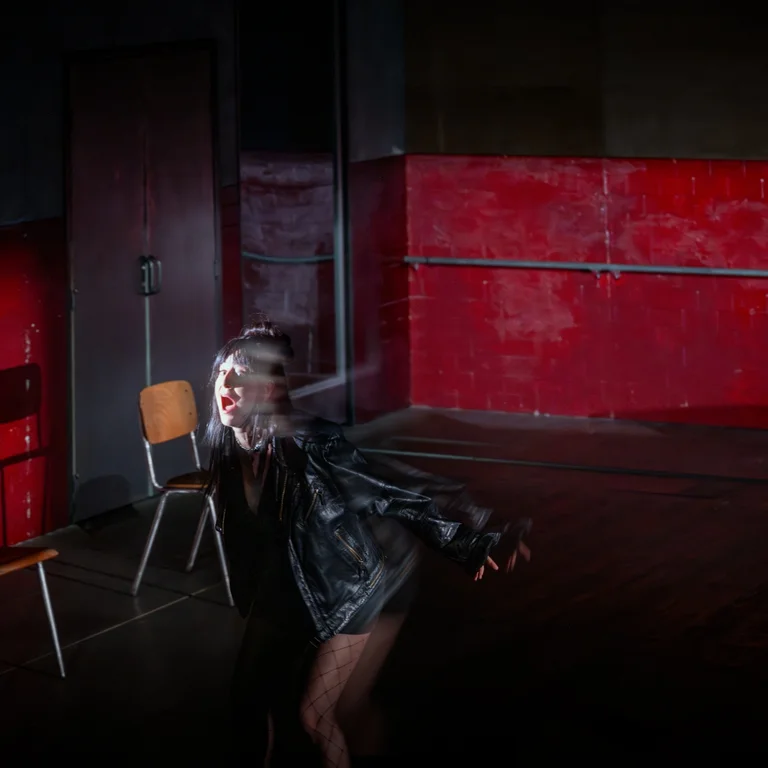 On a stage with red and grey wall surfaces, a person stands in the foreground with arms outstretched. Several chairs are arranged in the space, with focused lighting illuminating the scene.
