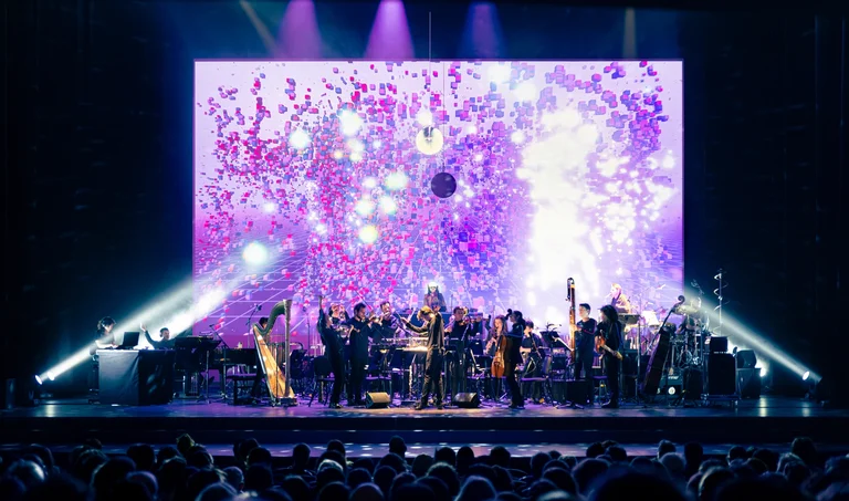 The orchestra Les Apaches ! performs for an audience, accompanied by a DJ artist and an impressive pink and purple digital light projection on stage.