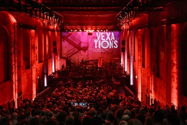 View across a densely seated audience in a large industrial hall towards the stage, where the ensemble Les Apaches ! is performing. The entire space is bathed in red light. The word “VEXATIONS” appears in large white letters on the back wall of the stage.