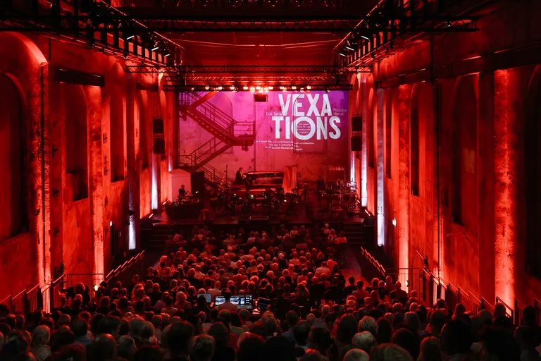 View across a densely seated audience in a large industrial hall towards the stage, where the ensemble Les Apaches ! is performing. The entire space is bathed in red light. The word “VEXATIONS” appears in large white letters on the back wall of the stage.