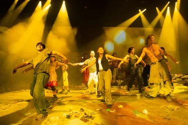A group of performers dances in unison on a stage. The focus is on the three performers at the front. The stage is covered in trash. Spotlights illuminate the scene in a smoky orange hue.