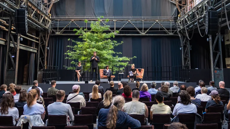 Discussion event in the Brave New Voices series at the Ruhrtriennale 2024 in the Jahrhunderthalle Bochum: Four people sit or stand on a stage in front of a large tree, surrounded by industrial architecture. In the foreground, an audience follows the discussion round attentively.