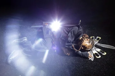 Scene from Balkan Erotic Epic: A person dressed in black lies on the ground surrounded by intense lens flare, with several silver blades arranged fan-like beside their head. The atmosphere feels dramatic and surreal.