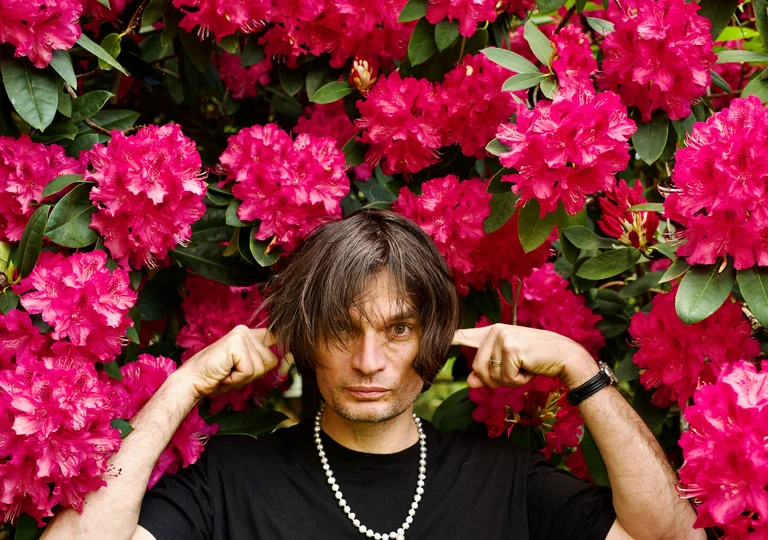 Jonny Greenwood stands in front of a background of vibrant pink blossoms. He is wearing a black T-shirt, a pearl necklace, and a wristwatch. With both hands, he covers his ears.