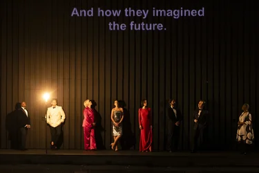 Schauspieler:innen stehen in einer Reihe vor einer dunklen Wand, während über ihnen die Worte "And how they imagined the future" projiziert werden.