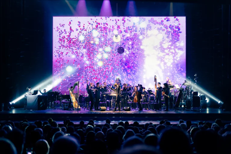 Les Apaches!  is performing on a stage in front of a glowing digital backdrop with purple and pink geometric shapes. Julien Masmondet stands at the center, while the musicians with string, wind, and percussion instruments, as well as a harp, form the ensemble.