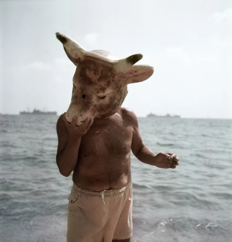 Ein Mann mit nacktem Oberkörper und hellen Badehosen steht knietief im Meer. Er trägt eine große Stierkopf-Maske, die sein Gesicht verdeckt. In einer Hand hält er eine Zigarette. Im Hintergrund sind mehrere Schiffe am Horizont zu sehen.