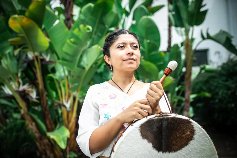 Nadia Larcher stands in front of large green leaves. She wears a white embroidered blouse and holds a large drum with a fur covering as well as a drumstick.