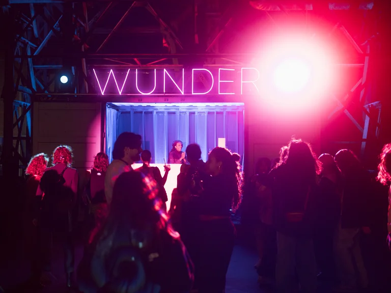 Eine Menschenmenge tanzt vor einem blau beleuchteten Container vor einer dunklen Industriehalle. Über dem Container leuchtet der Schriftzug „WUNDER“ in violettem Neonlicht, während ein intensiver pink-roter Scheinwerfer die Szene erhellt.