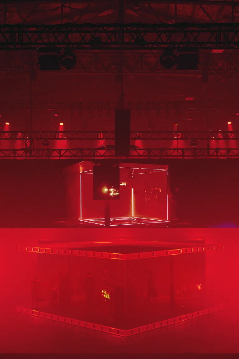 Stage scene in deep red lighting with a cube-shaped structure and silhouetted figures.