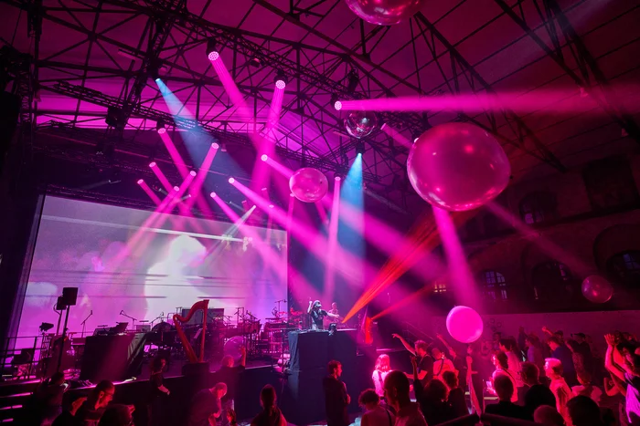 In alten Gladbecker Maschinenhalle tanzt ein Publikum ausgelassen vor einer Bühne, auf der ein DJ und Musiker:innen live performen. Über der Tanzfläche schweben große transparente Bälle, während ein Lichtdesign mit pinken und blauen Strahlen die Szene in ein futuristisches Ambiente taucht.