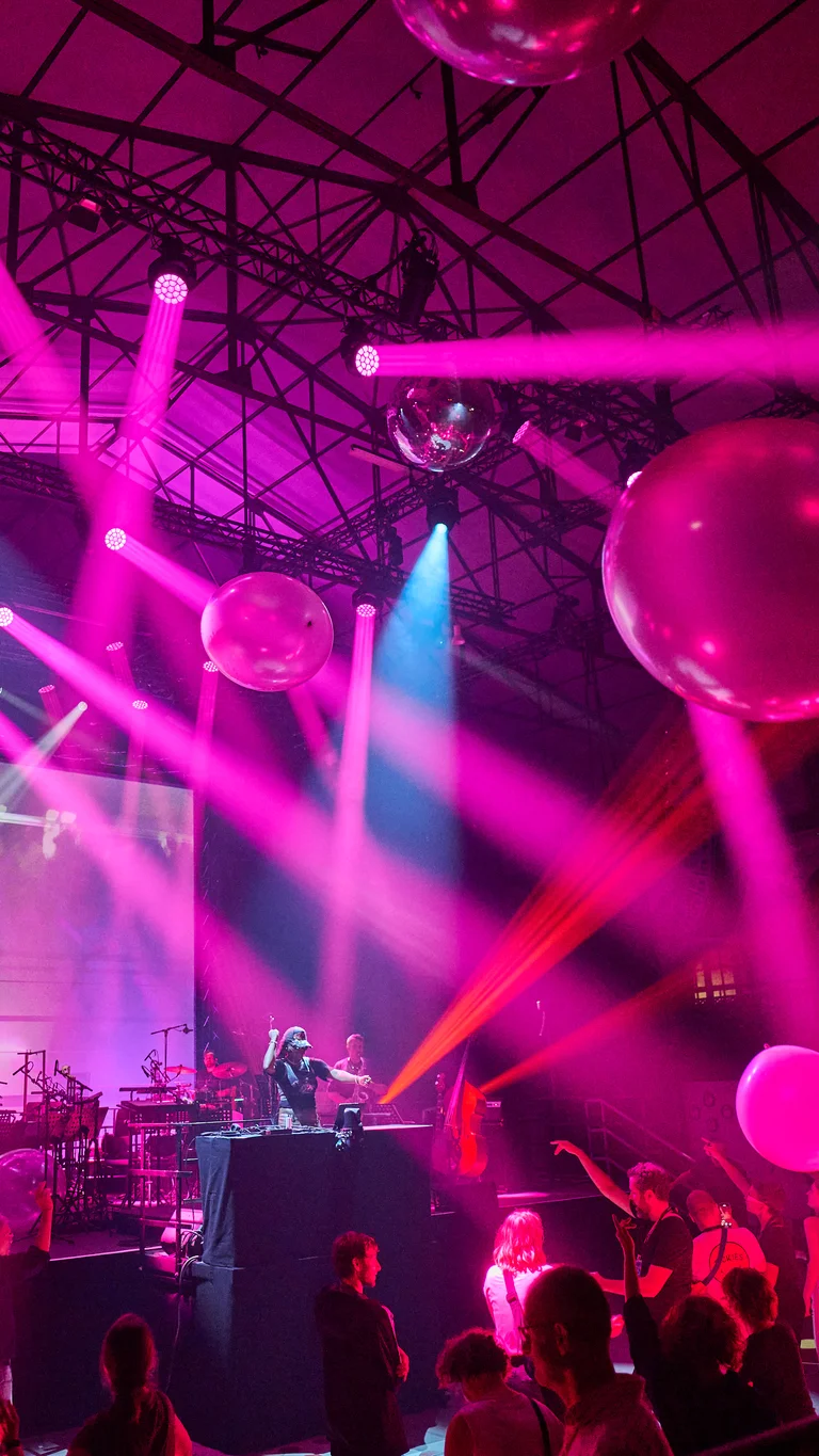 In the old machine hall in Gladbeck, a crowd dances in front of a stage where a DJ and musicians perform live. Large transparent balloons float above the dance floor, while pink and blue lighting design bathes the scene in a futuristic atmosphere.