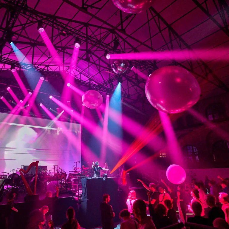 In the old machine hall in Gladbeck, a crowd dances in front of a stage where a DJ and musicians perform live. Large transparent balloons float above the dance floor, while pink and blue lighting design bathes the scene in a futuristic atmosphere.