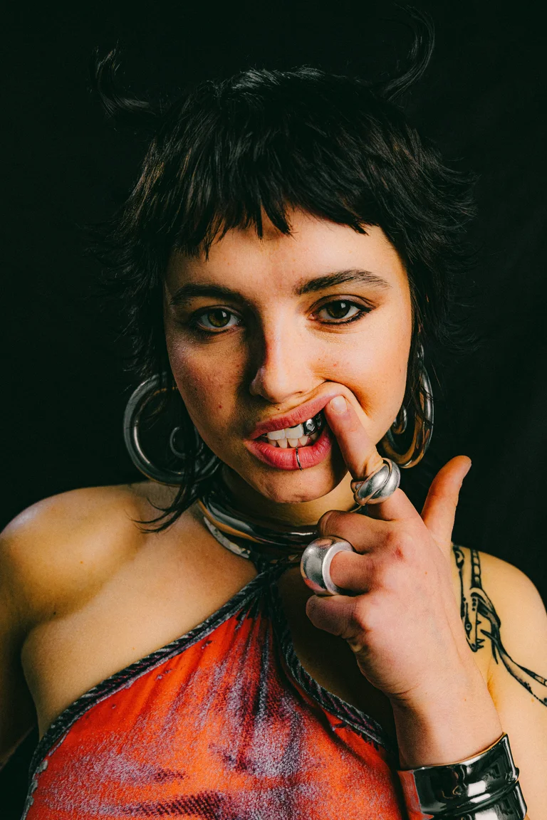 The artist Marnele looks directly into the camera against a black background. She has short black hair, styled into small horns on the sides. With one finger, she pushes her upper lip upwards, revealing her grillz on her teeth.