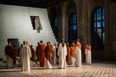 A group of performers in white and orange robes stands before a monumental, pale staircase structure. Above them, a performer in a shimmering gold robe stands in an opening in the set.