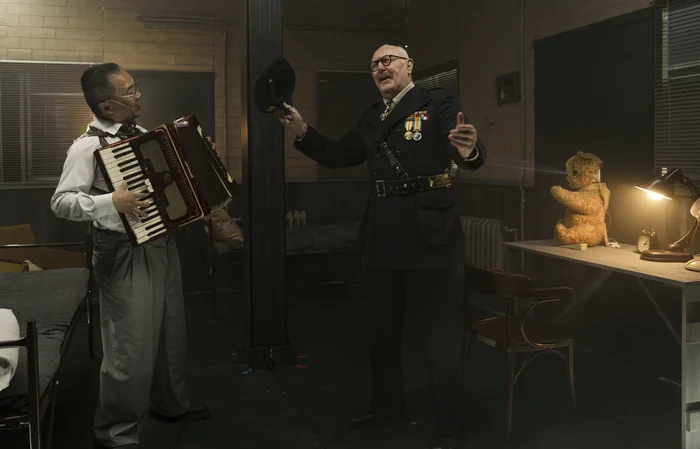 Zwei Darsteller auf der Bühne; einer spielt Akkordeon, der andere trägt eine Uniform mit Orden und hebt den Hut.