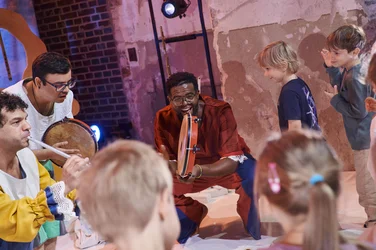 Drei Musiker spielen Flöte und Trommeln inmitten einer Gruppe von Kindern.