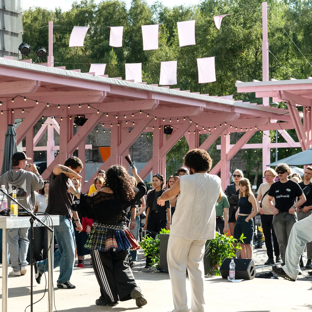 Die Mitglieder von Patina Records unter der rosafarbenen Holzkonstruktion des Festivalzentrums Wunderland; davor Publikum, das die Show verfolgt.