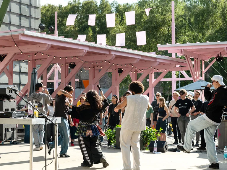 Die Mitglieder von Patina Records unter der rosafarbenen Holzkonstruktion des Festivalzentrums Wunderland; davor Publikum, das die Show verfolgt.