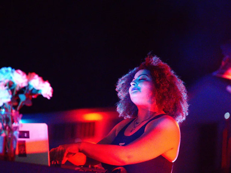 DJ Gîn Bali performs at a booth under colorful club lighting, with a vase of flowers beside her.