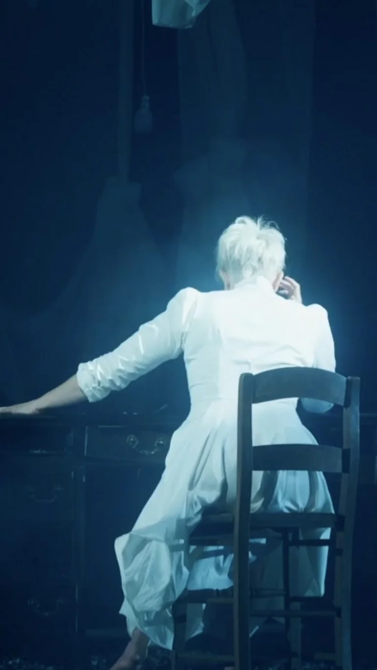Joyce DiDonato sitzt in einem weißen Kleid an einem Schreibtisch, den Rücken zur Kamera gewendet.
