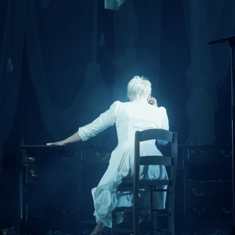 Joyce DiDonato sitzt in einem weißen Kleid an einem Schreibtisch, den Rücken zur Kamera gewendet.