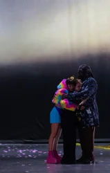 Three people stand closely embracing on a stage. One person wears a colorful fluffy jacket and pink platform boots, while another wears a dark coat. Pink flower petals are scattered across the floor.
