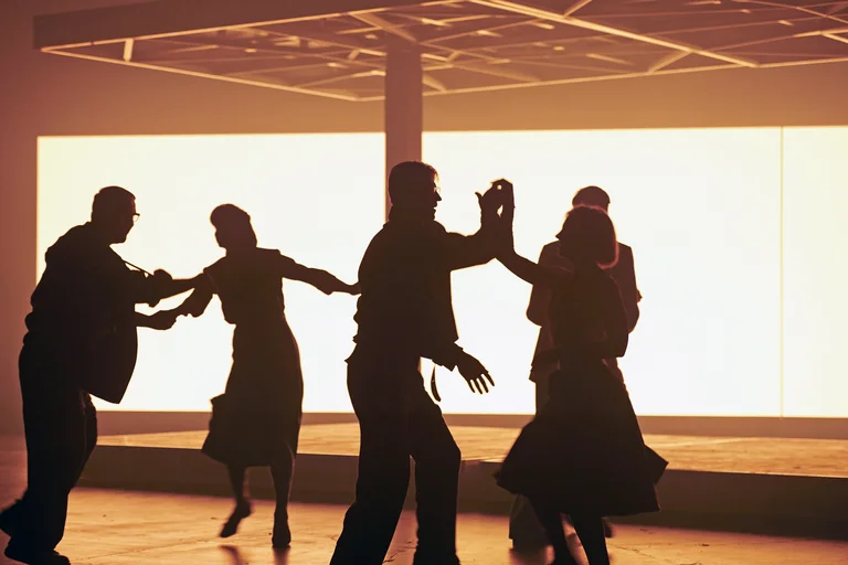 Dance scene on stage: Silhouettes of several actors against a brightly lit background.