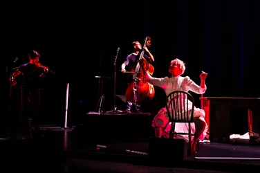 Joyce DiDonato sits on a chair in a white dress, raising both arms. Behind her, Charles Yang plays the violin and Ranaan Meyer the double bass. The dark stage is illuminated by red light.