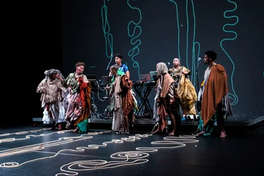 Mehrere Performer:innen in kunstvollen Kostümen auf einer Bühne, die sich in ausdrucksstarken Posen bewegen. Vor ihnen auf dem Boden liegen Seile kunstvoll angeordnet. Diese Anordnungen finden sich im Hintergrund als Projektion wieder.