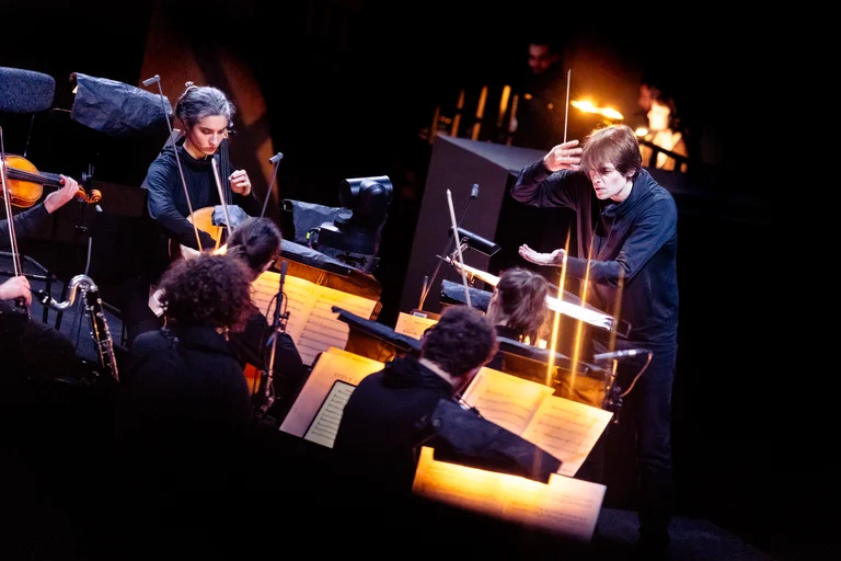 Julien Masmondet conducts an orchestra on a dark stage. He raises the baton with a focused expression while the musicians, surrounded by warmly lit music stands, play their instruments. In the foreground, string and wind players can be seen.