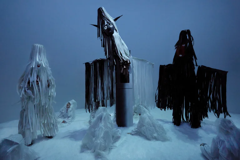 Blue-lit snowy scene with three tall fringed figures in white and black, while several white-clad people kneel on the ground.