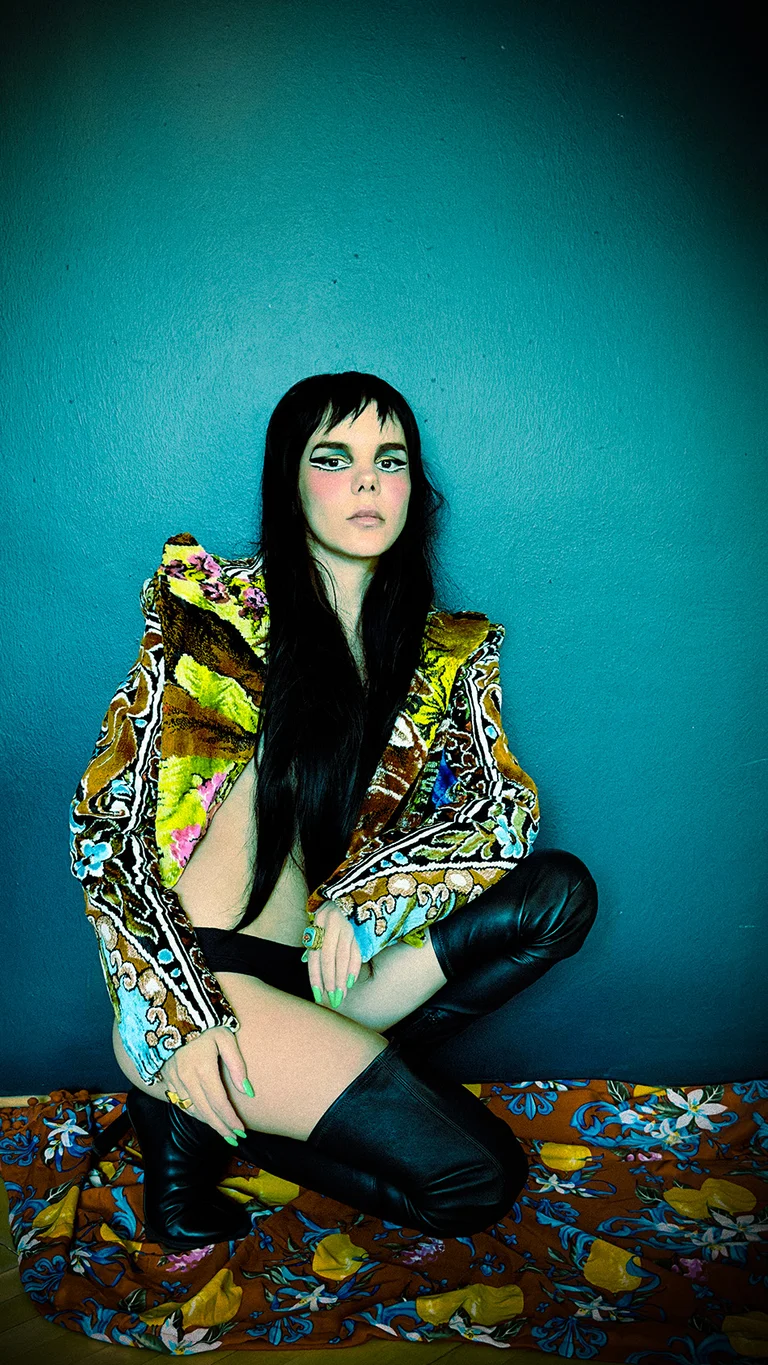 The singer Gaye Su Akyol sits in front of a teal-colored wall. She wears a patterned, colorful jacket, black over-the-knee boots, and sits on a vibrant floral-patterned cloth. The lighting creates a dramatic contrast against the dark background.