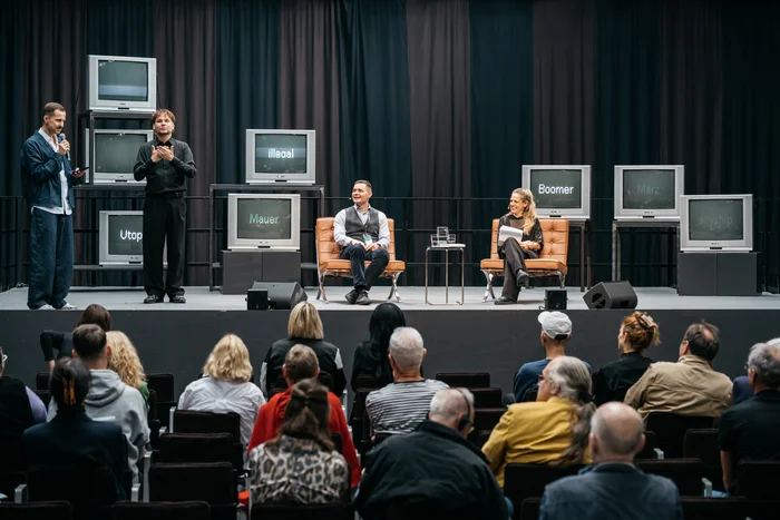 Auf einer Bühne sitzen Dr. Janosch Schobin und Sivan Ben Yishai in Sesseln im Gespräch. Links von ihnen steht ein Mann mit Mikrofon, daneben ein Gebärdensprachdolmetscher. Mehrere alte Fernseher im Hintergrund zeigen Wörter wie „illegal“, „Mauer“, „Utopie“, „Boomer“, „März“ und „allyship“. Im Vordergrund ist das Publikum zu sehen.