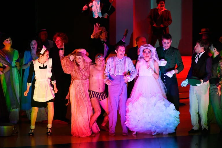 A group of people in costumes, including a chambermaid and a bride, stand on a colourfully lit stage.