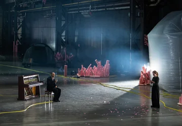 Two people are on a spacious stage in an industrial hall. One person sits beside an open upright piano, while the other stands opposite holding a sheet of paper. Pink foam elements, yellow cables, and scattered flower petals cover the floor; a tent and a large inflatable structure are visible in the background.
