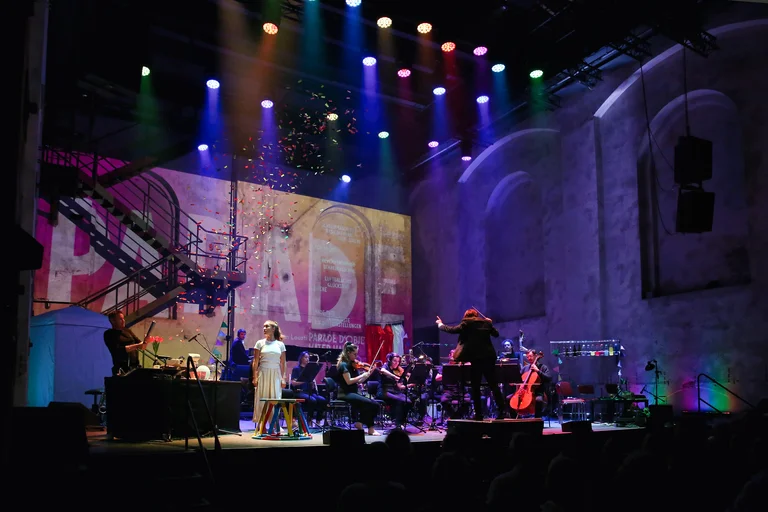 The ensemble Les Apaches ! performs on stage in an industrial hall, while Sarah Silverblatt-Buser stands to the left of the musicians. A conductor leads the ensemble. Colourful spotlights illuminate the stage as multicoloured confetti falls from above.