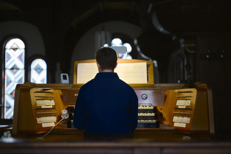 Organist James McVinnie seated at the organ console in the Evangelical Church at the Market in Katernberg during the event 124 Years of Reverb. Seen from behind, he plays at the illuminated keyboard with the stops and sheet music clearly visible. Stained-glass windows shimmer faintly in the background.